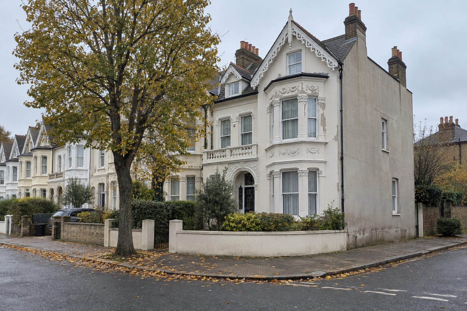 A large cream stucco Victorian villa with ornate bay windows and an arched doorway on a street corner, beside a tree in autumn leaf