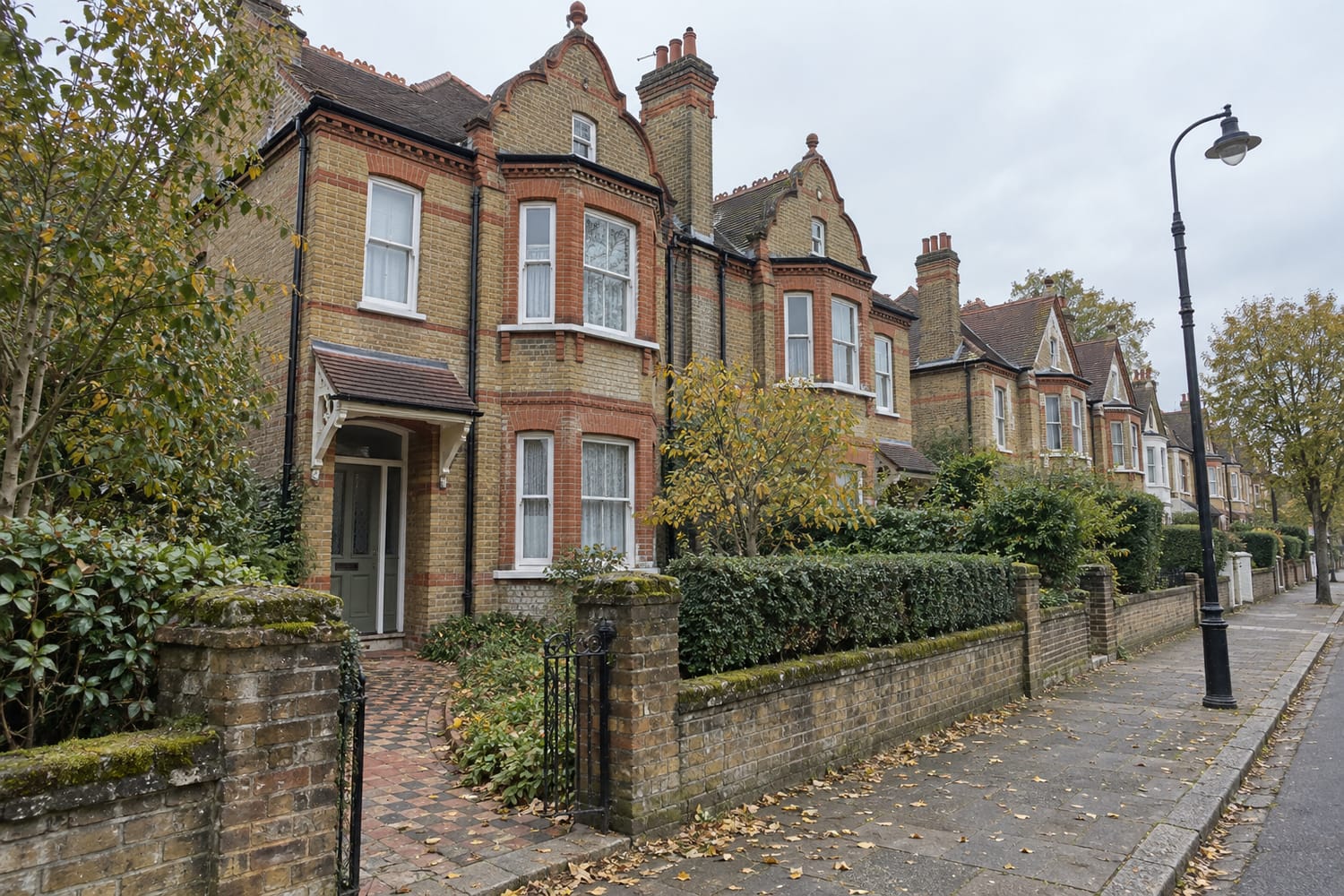 Yellow brick semi-detached houses with red brick bands and curved gables behind a low wall, beside an old street lamp