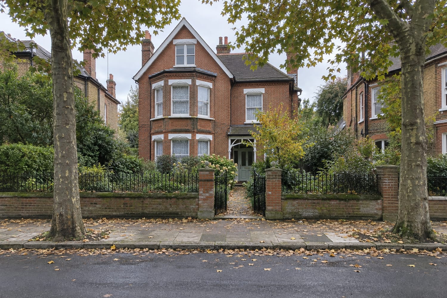 A detached red brick Victorian house set back behind railings and a front garden, framed by two plane trees